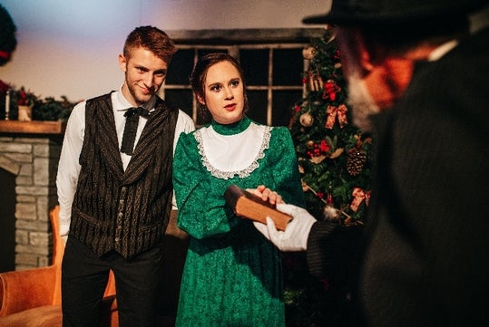 A woman in a green dress and a man in a vest stand indoors near a decorated Christmas tree, facing an older man holding out a wooden object.