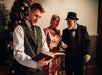 Three people in Victorian-style clothing stand indoors by a decorated Christmas tree; one man in front is reading an open book and two others are conversing in the background.
