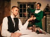A man in period clothing sits at a table holding papers while a woman in a green dress decorates a small Christmas tree in a vintage-styled room.