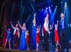 Performers on stage wearing formal attire, raising their arms in unison during a lively musical number with a colorful backdrop.