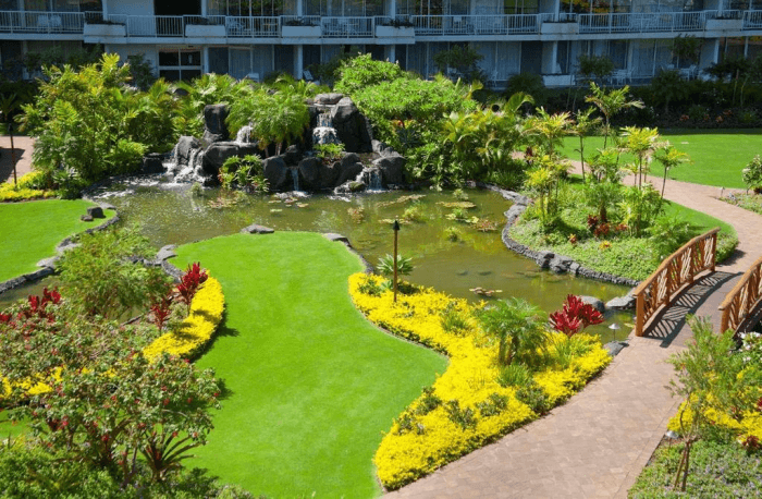 A landscaped garden with a pond, rocks, a waterfall, and a wooden bridge, surrounded by pathways and lush greenery.