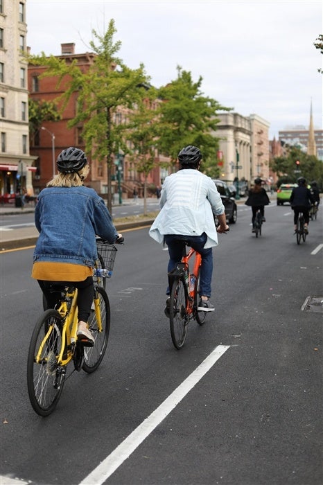People riding bicycles on a city street with helmets. Trees and buildings line the road.
