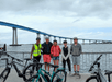 Five people with bicycles stand on a waterfront pier, wearing helmets and casual clothing. A large bridge spans the background under a cloudy sky.