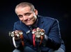 Man in a blue blazer holding multiple wristwatches in both hands against a dark background.