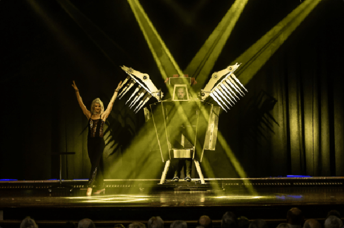 A woman in a black dress with her arms up on stage next to a man in an elaborate box with yellow light shining on them.
