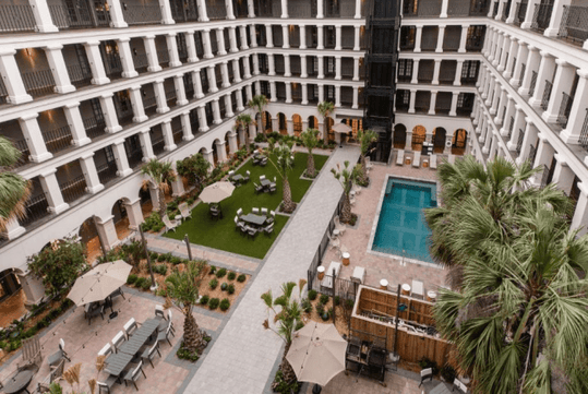 A large hotel courtyard with a central swimming pool, green lawn area, outdoor seating, palm trees, and surrounding multi-story balconies.