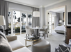 Modern hotel suite with a living area featuring a sofa, round table, and a TV. Bedroom visible through open doors. Large windows with curtains overlook palm trees. Neutral color scheme.
