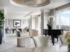 Luxurious living room with modern decor, featuring a grand piano, abstract artwork, and a chandelier. Large windows provide a view of hills. Neutral tones dominate the space.