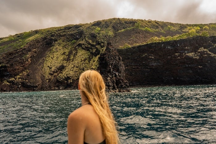 A person with long blonde hair stands on a boat, looking at a rugged, green-covered cliff above blue ocean water. Cloudy sky above.