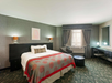 Hotel room with a large bed, decorative bedspread, wall art, two lamps, a window, desk, chair, and side table on carpeted floor.