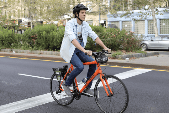 A person wearing a helmet rides an orange bicycle on the road, crossing a white pedestrian line.