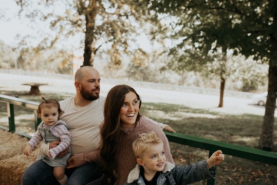 A family of four sits outdoors on hay bales, with trees and a road in the background. The adults and two children appear to be enjoying the day.