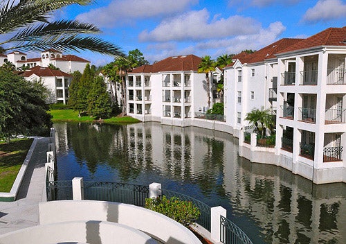 White apartment buildings with red roofs surround a calm canal. Palm trees and lush greenery line the water's edge under a partly cloudy sky.