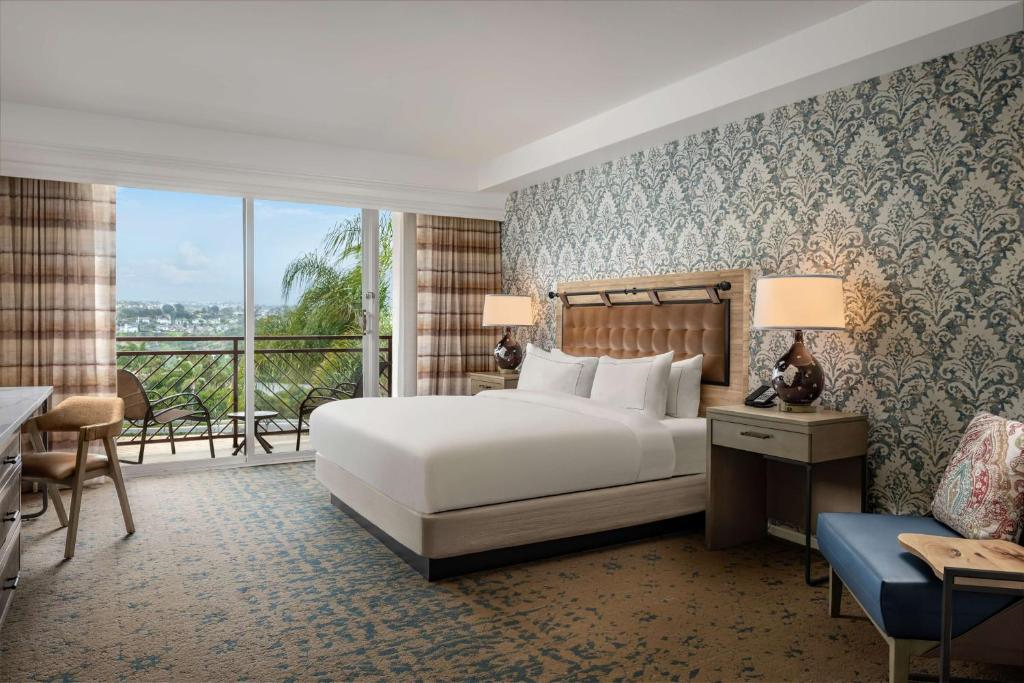 Hotel room with a large bed, patterned wallpaper, two bedside lamps, a window with curtains, and two chairs by a table.