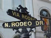 Street signs for Via Rodeo and N. Rodeo Dr intersect against a backdrop of a building with an ornate window.