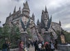 People walk toward a large, castle-like structure with spires and stone walls. Two winged statues flank the entrance. Overcast sky in the background.