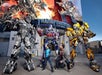 Two people pose with large Transformer robot characters outside "Transformers: The Ride" attraction.