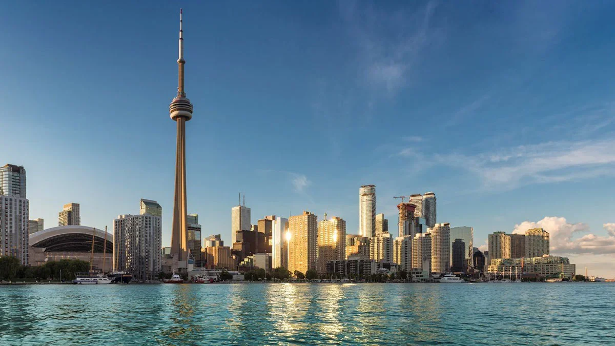 The Toronto skyline at sunrise, featuring the CN Tower and waterfront buildings reflecting in the calm water.