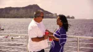 couple holding drinks at Spirit of Aloha Catamaran Sunset Sail with water in background and mountain and buildings in distance in Big Island, Hawaii, USA